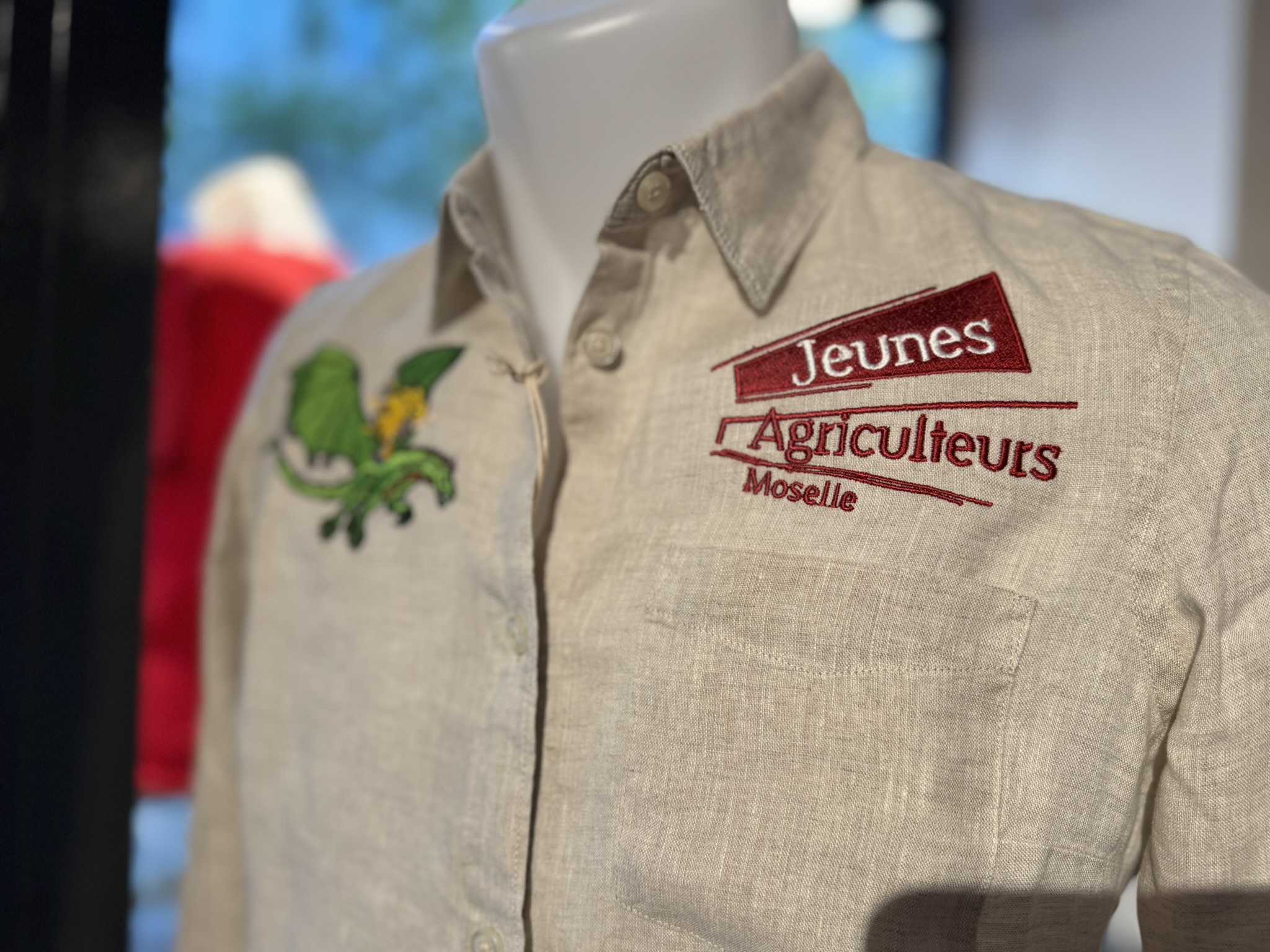 Chemise beige en lin portée sur un mannequin, brodée des inscriptions « Jeunes Agriculteurs Moselle » en rouge, avec un motif végétal vert et jaune sur la poitrine gauche.