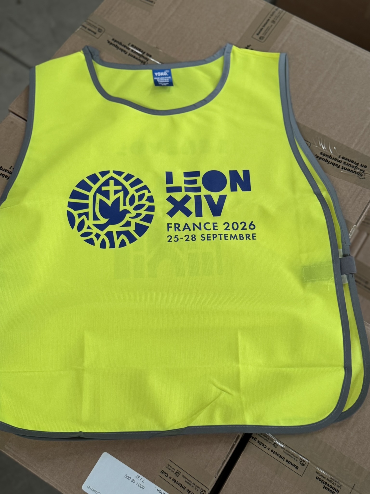 Gilet de sécurité jaune fluorescent « LÉON XIV – FRANCE 2026 – 25-28 SEPTEMBRE », posé sur des cartons.